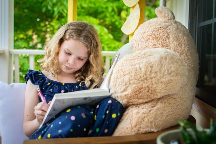 Young blond girl reads next to teddy bear
