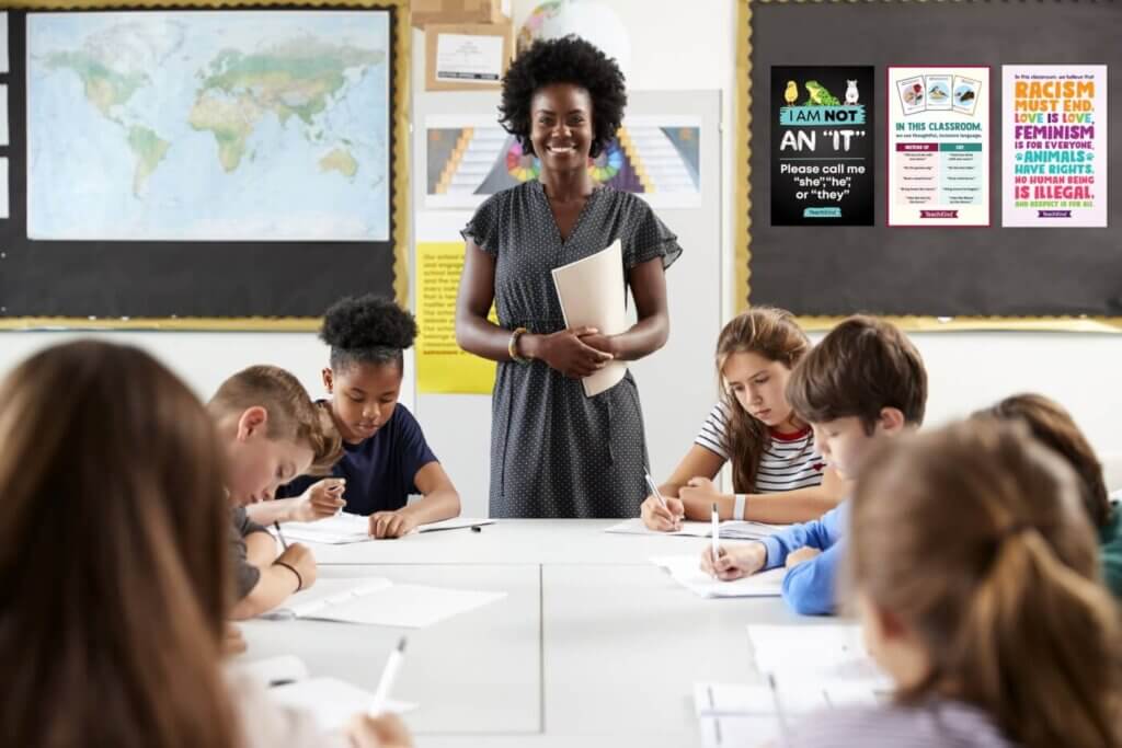 A teacher is standing at the front of a classroom while elementary school students do an activity at a table. 