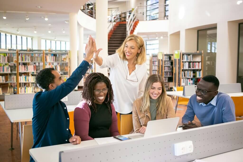 Cheerful teachers gathered around a computer in a library, two of whom were having a high five