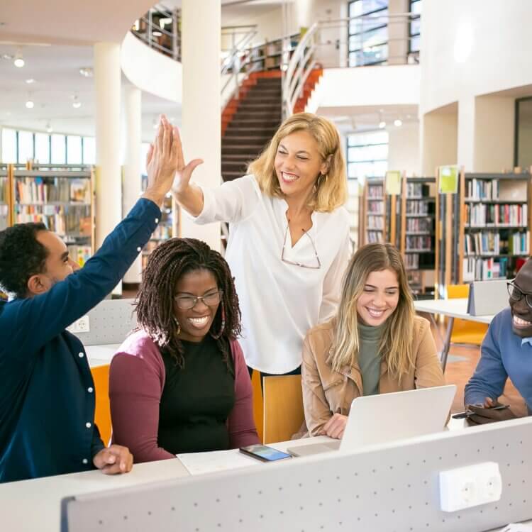 Cheerful teachers gathered around a computer in a library, two of whom were having a high five