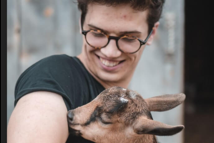 A young person smiles as a baby goat (kid) nuzzles their arm