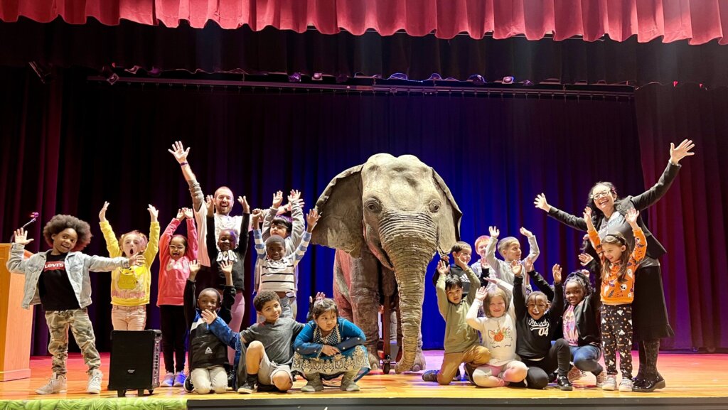 Students celebrate with their hands in the air next to Ellie, the animatronic elephant
