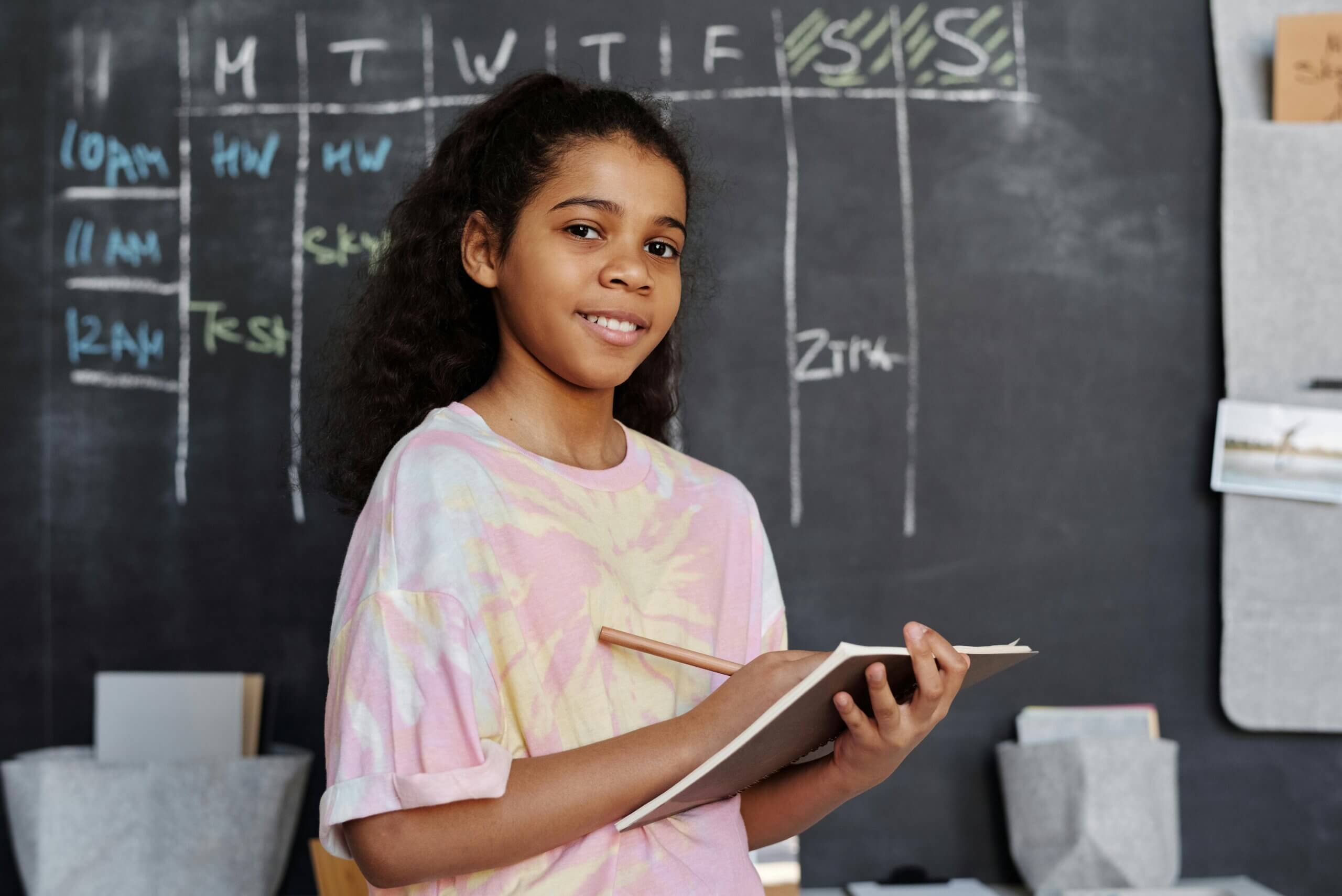Gril in Pink Crew Neck T-shirt Holding Notepad in front of a chalkboard