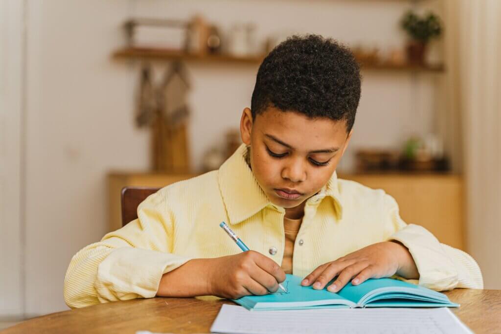 A young student with a yellow shirt focused on writing in a notebook.