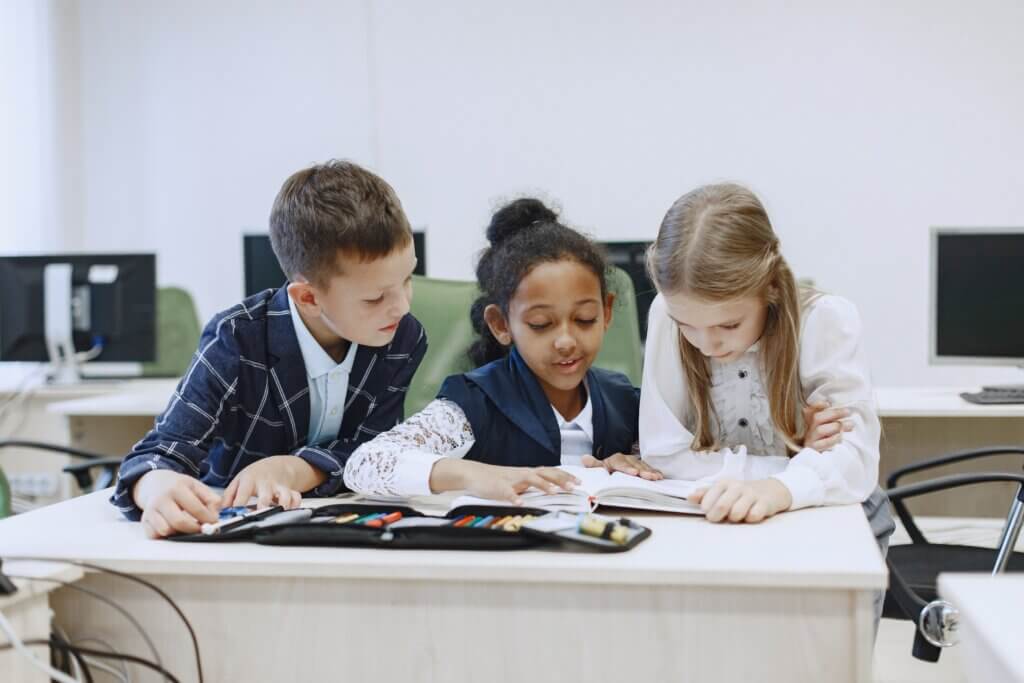 Three children working together at a desk in a classroom, focusing on a book.