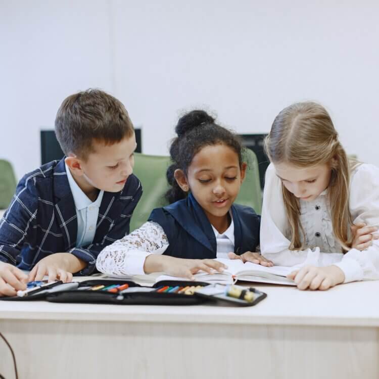 Three children working together at a desk in a classroom, focusing on a book.