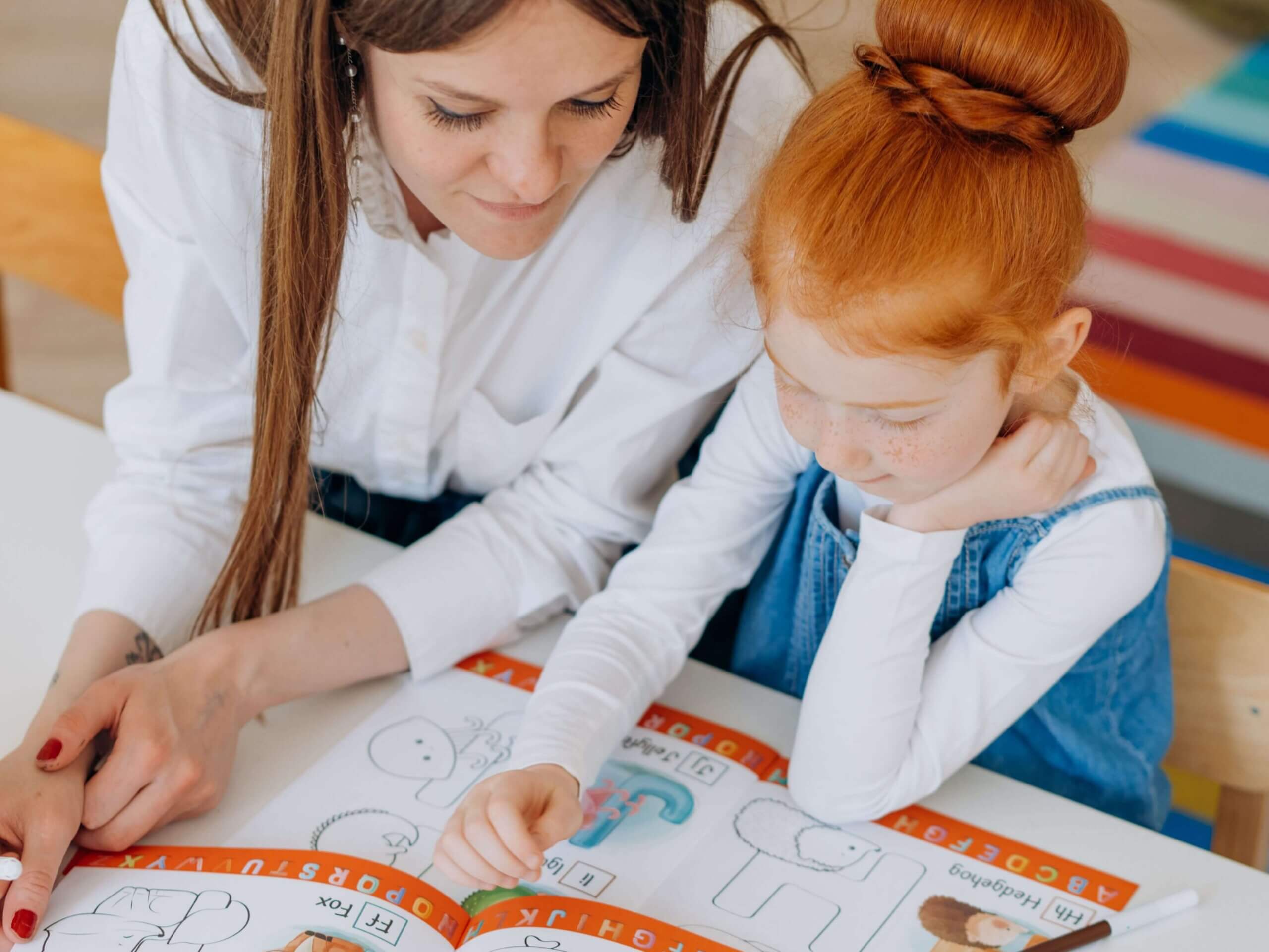 A teacher helping a young girl with letter recognition in a colorful classroom setting.