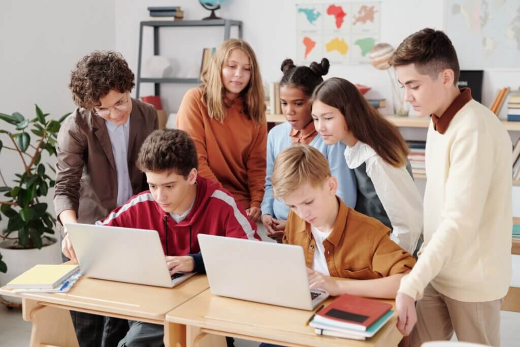 A group of middle school students collaborating on laptops in a classroom setting, guided by a teacher.