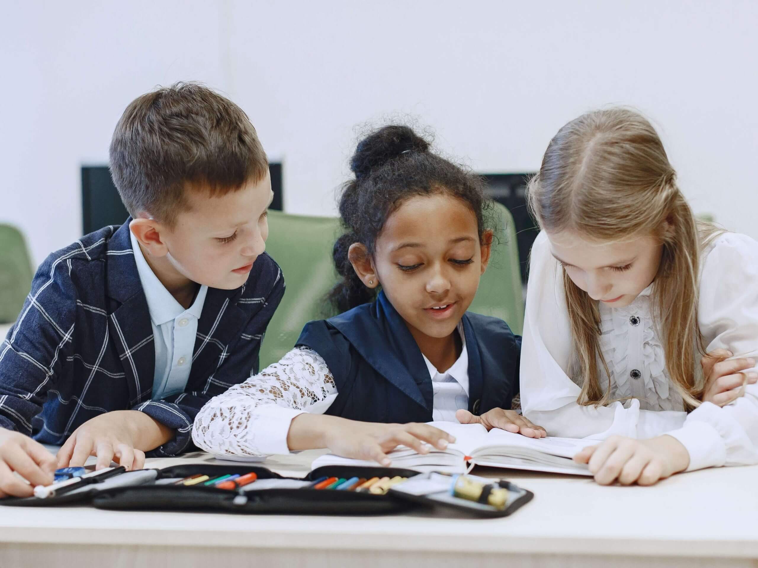 Three children working together at a desk in a classroom, focusing on a book.