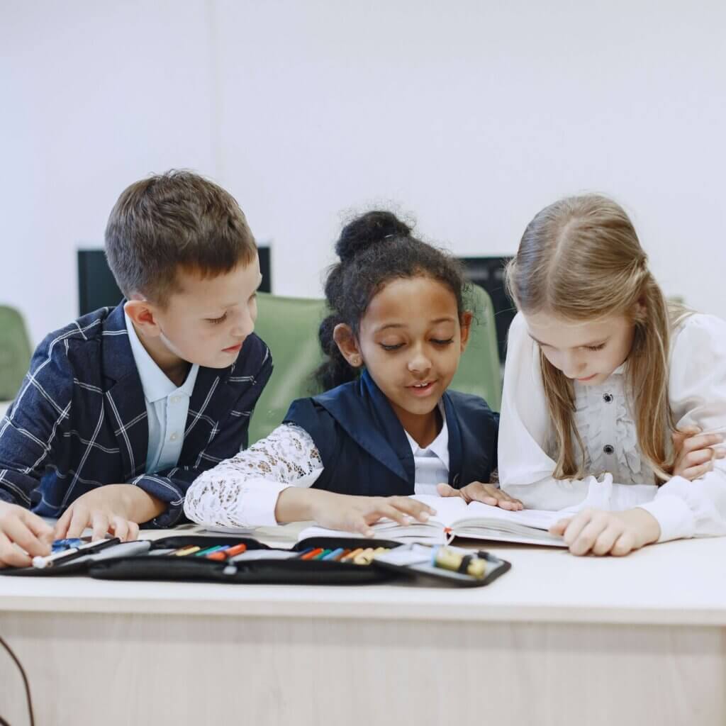 Three children working together at a desk in a classroom, focusing on a book.